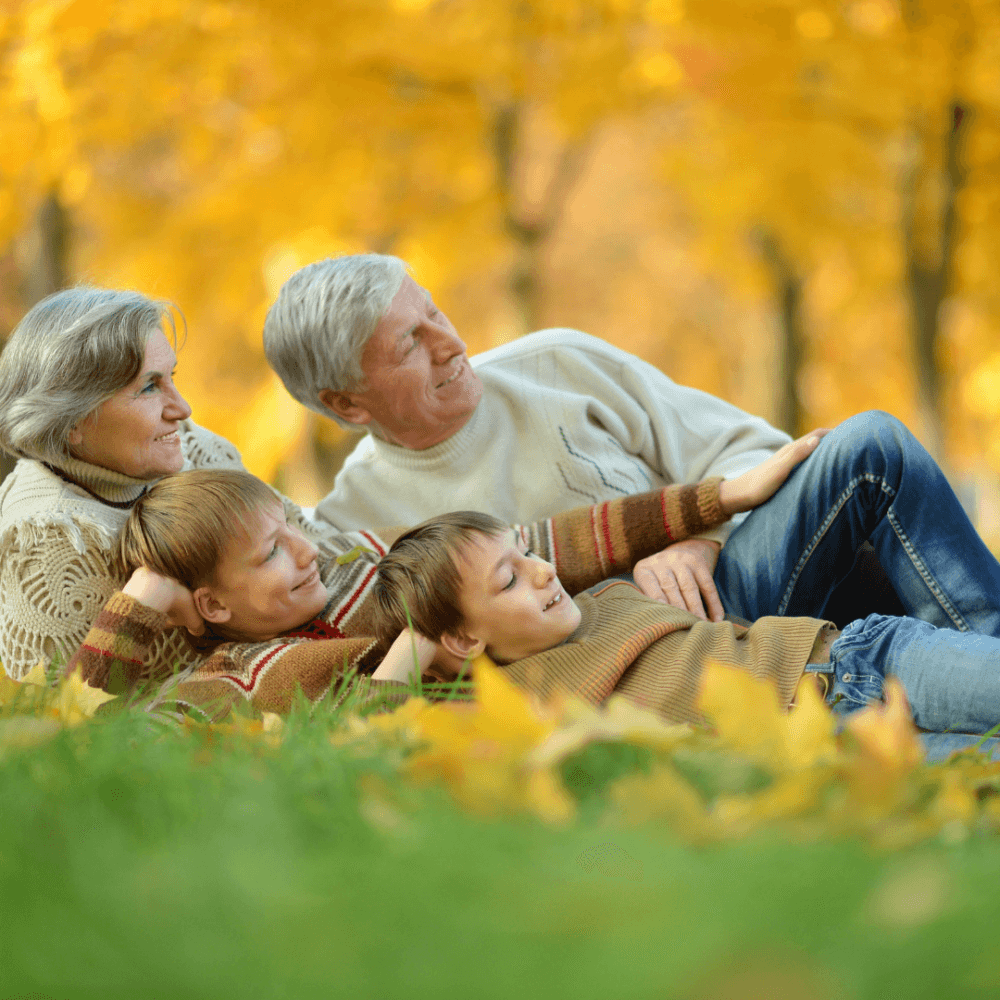 A smiling elderly couple and two young boys lie on grass in a park surrounded by vibrant yellow autumn leaves. - Home Instead