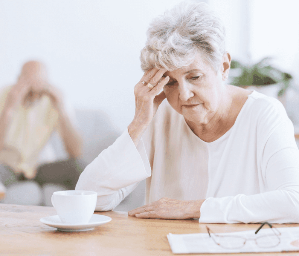 Elderly woman sitting at a table, looking worried, with a cup of coffee, holding her head. An elderly man is in the background. - Home Instead
