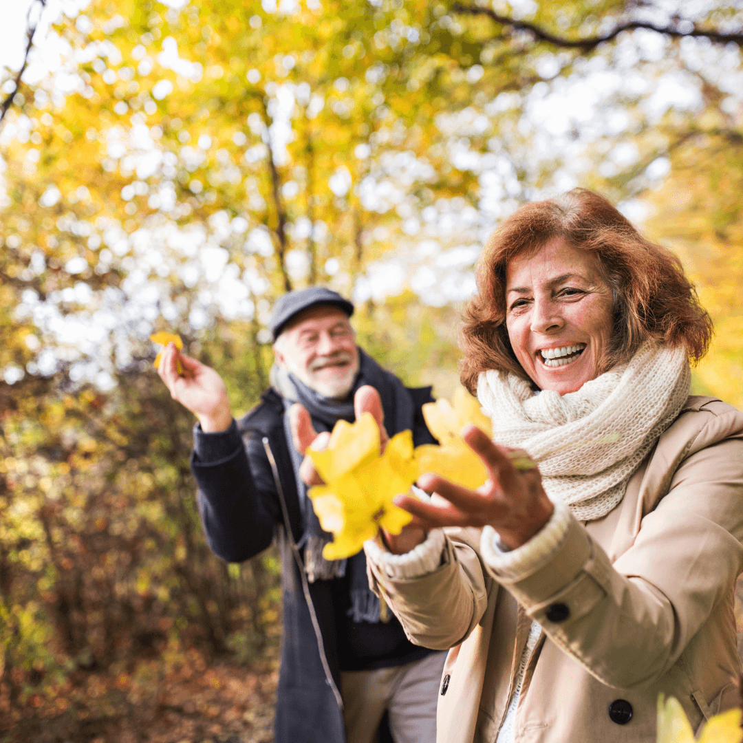 Elderly couple enjoying a walk in the autumn woods, smiling and holding yellow leaves. - Home Instead