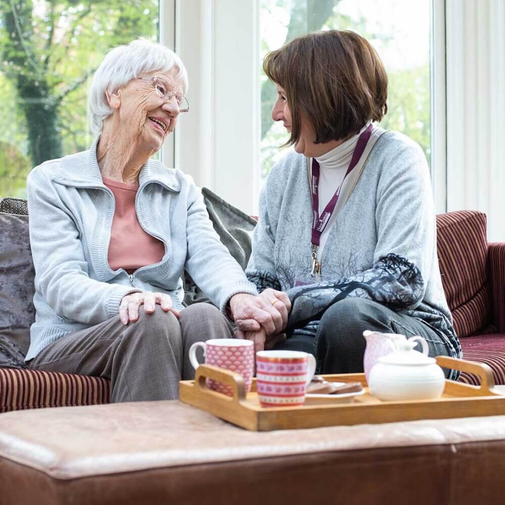An elderly woman and a younger woman sit on a couch, holding hands and smiling, with tea cups on a tray in front of them. - Home Instead