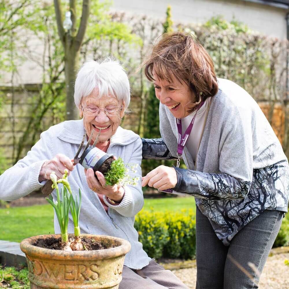 Two women smiling and gardening together, one holding a small potted plant and scissors. - Home Instead