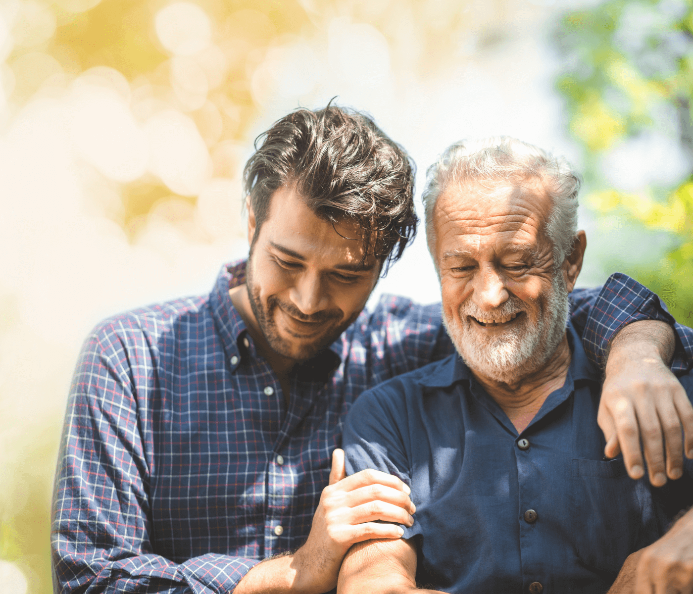 Younger person and older person smiling and embracing each other in a sunlit, outdoor setting. - Home Instead