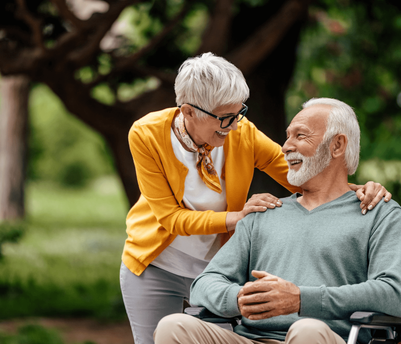 Elderly woman with short gray hair and yellow sweater smiles at elderly man with white beard in a wheelchair outdoors. - Home Instead