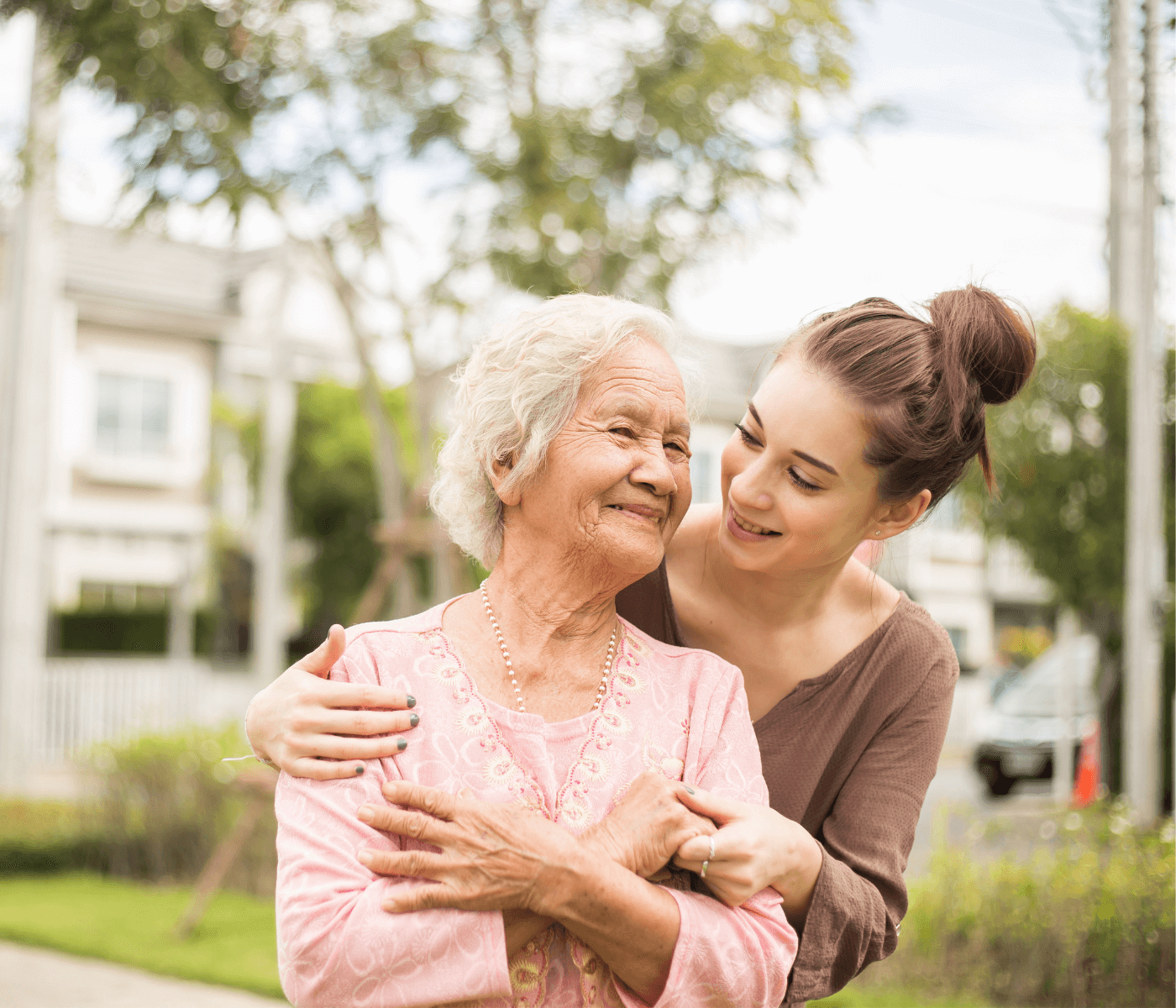 A young woman embraces an elderly woman outside, both smiling warmly at each other. - Home Instead