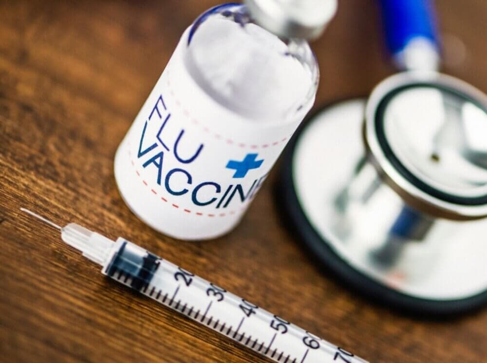 Flu vaccine vial, syringe, and stethoscope on a wooden surface, representing medical tools for flu prevention. - Home Instead