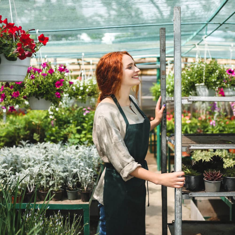A woman in an apron smiles while arranging plants on a metal rack in a greenhouse filled with various flowers. - Home Instead