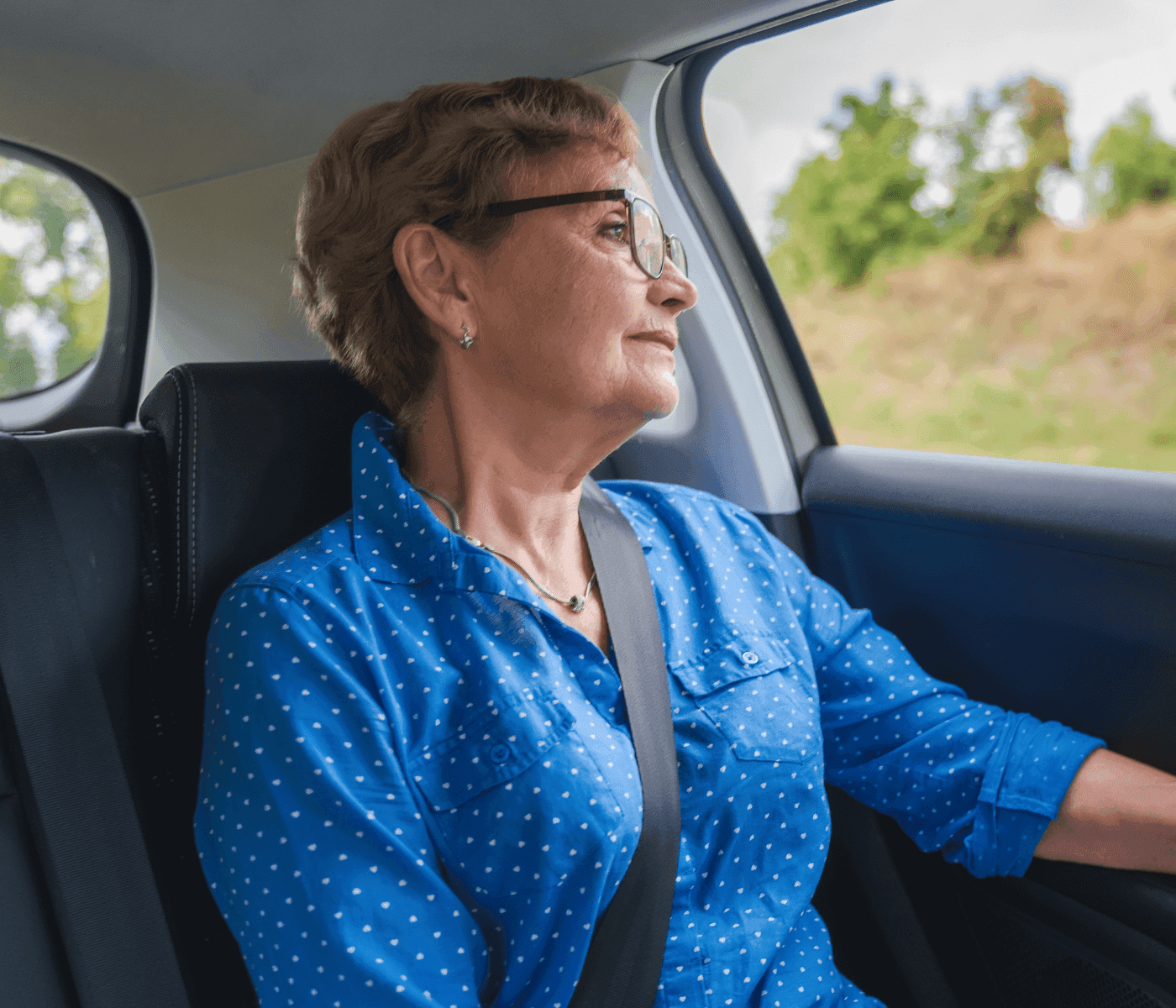 Elderly woman wearing glasses and a blue polka-dot shirt, seated in a car and looking out the window with a thoughtful expression. - Home Instead