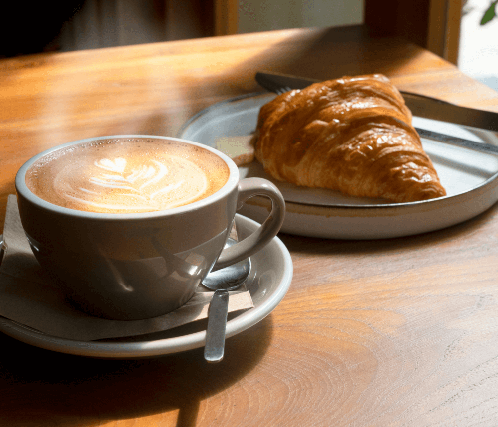 A cup of latte with latte art and a croissant on a plate, placed on a wooden table in sunlight. - Home Instead