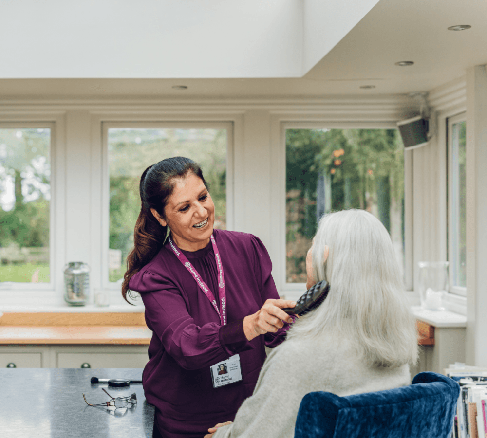 A caregiver in a purple top smiles while brushing a seated elderly woman's hair in a bright room. - Home Instead