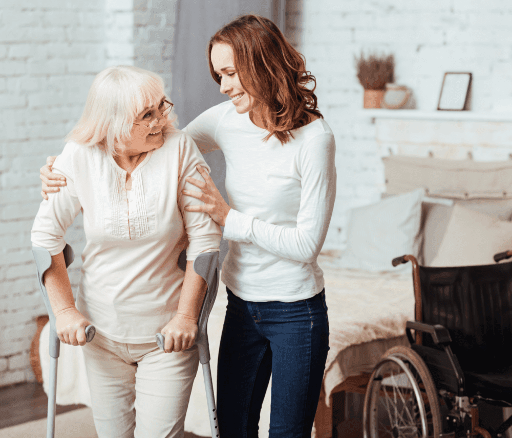 A young woman supports an elderly woman with crutches, smiling together in a living room, with a wheelchair nearby. - Home Instead