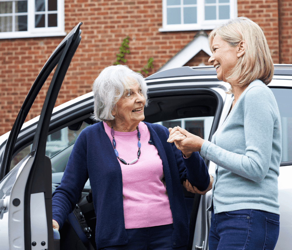 Two women stand by a car, one older woman with gray hair is holding the hand of a smiling younger woman. - Home Instead
