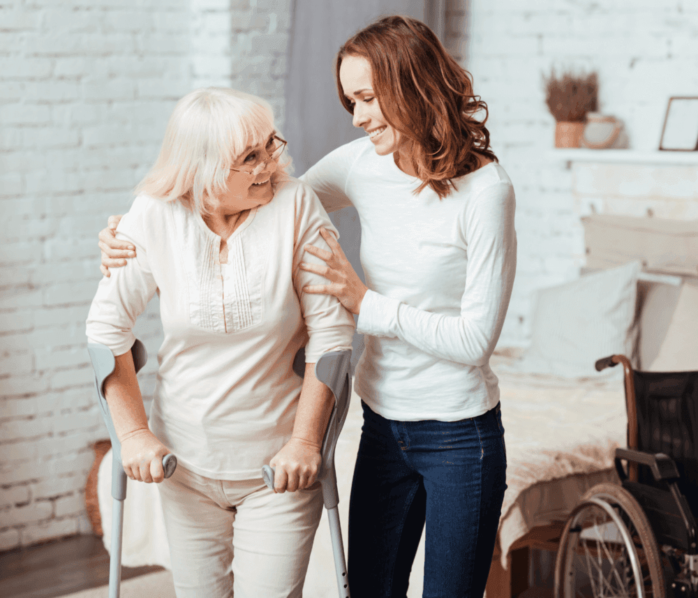 A young woman smiling and supporting an elderly woman using crutches in a cozy, well-lit living room with a wheelchair nearby. - Home Instead