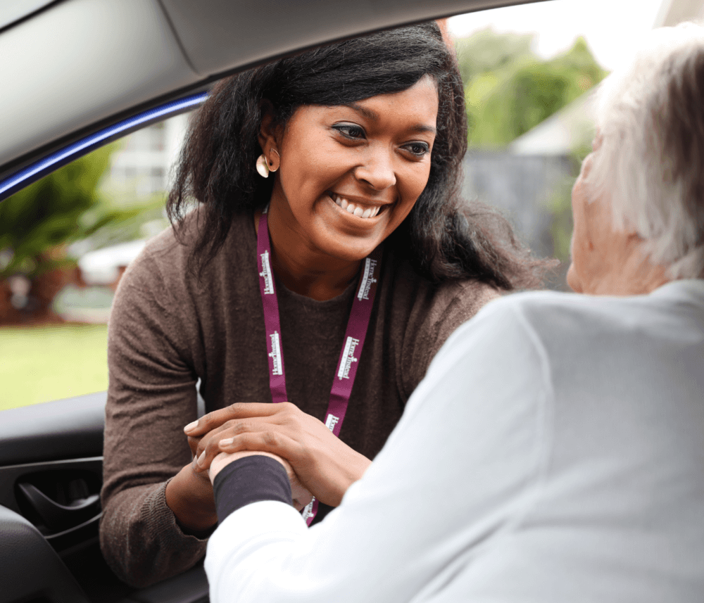 A smiling woman with a lanyard helps a seated elderly person inside a car. - Home Instead
