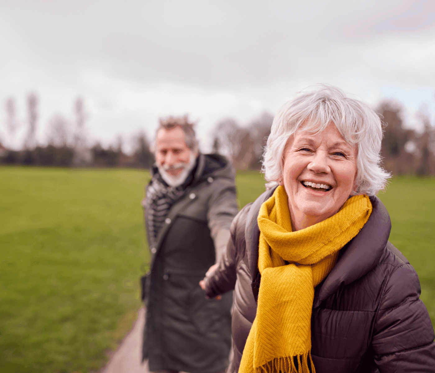 Elderly couple smiling, holding hands, while walking in a park with greenery in the background. - Home Instead