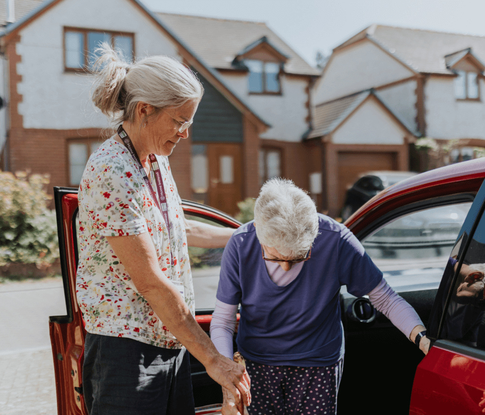 A woman helps an elderly person out of a red car in a residential neighborhood. - Home Instead