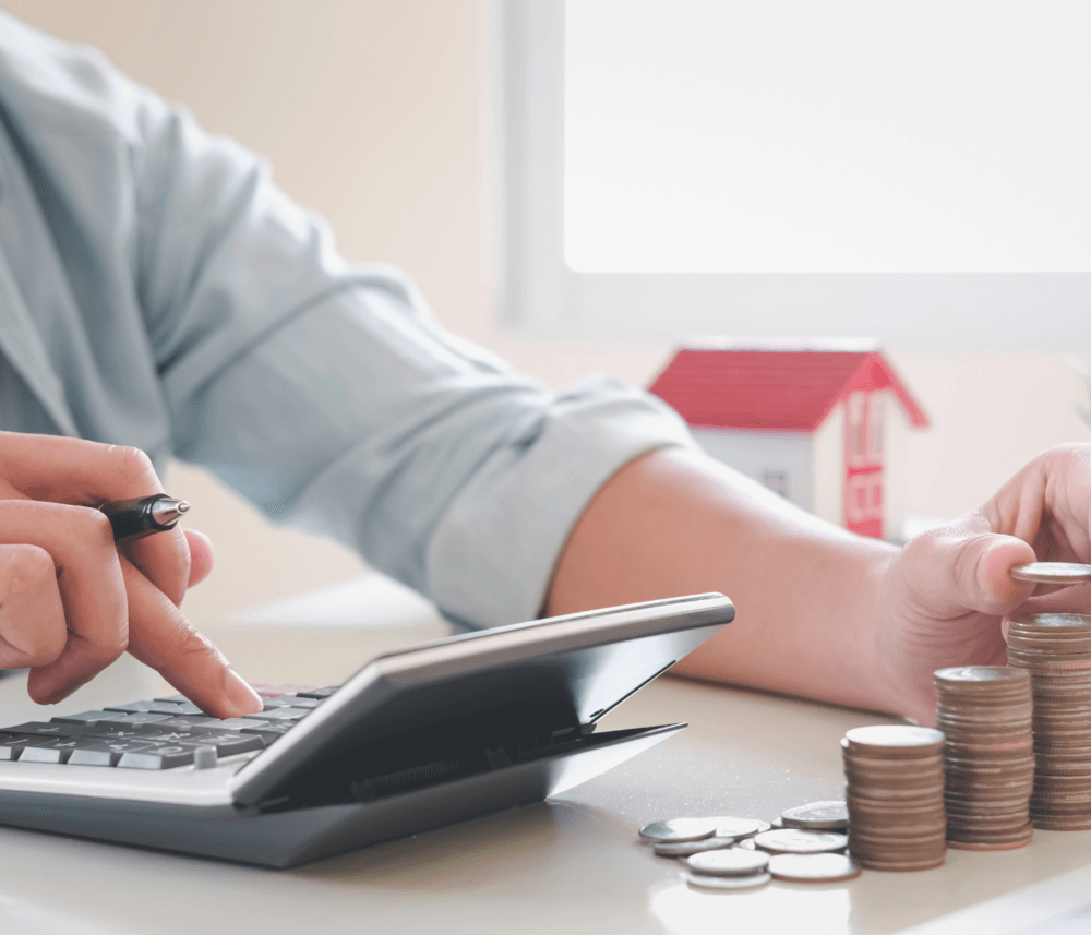 Person using a calculator with one hand and stacking coins with the other, small house model in the background. - Home Instead