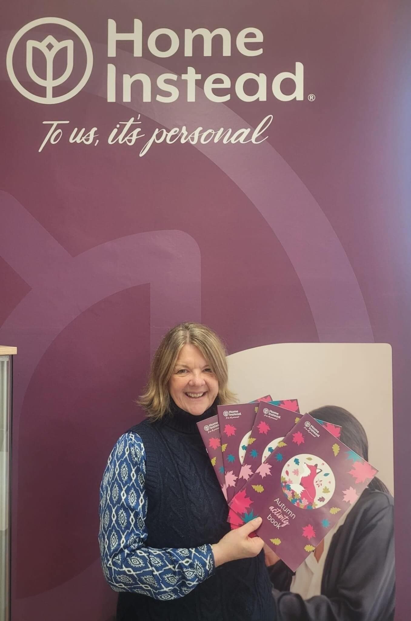 Smiling person holding booklets in front of a purple "Home Instead" sign with the tagline "To us, it's personal. - Home Instead