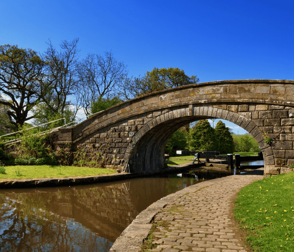 A stone bridge arches over a serene canal beside a cobblestone path, surrounded by lush greenery and trees under a blue sky. - Home Instead
