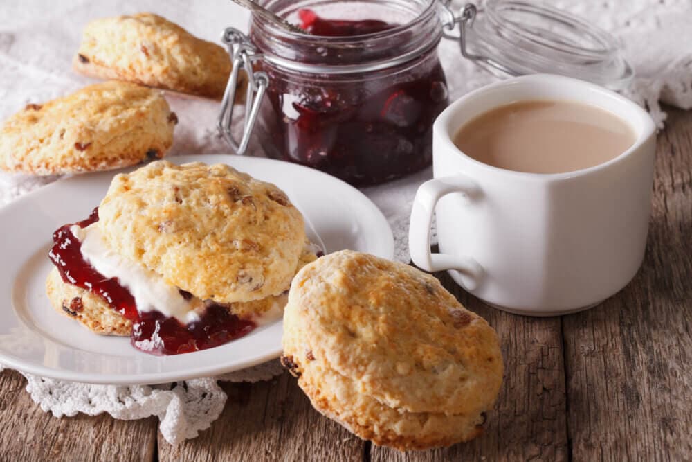 Scones with cream and jam, a jar of jam, and a cup of tea with milk on a rustic wooden table. - Home Instead