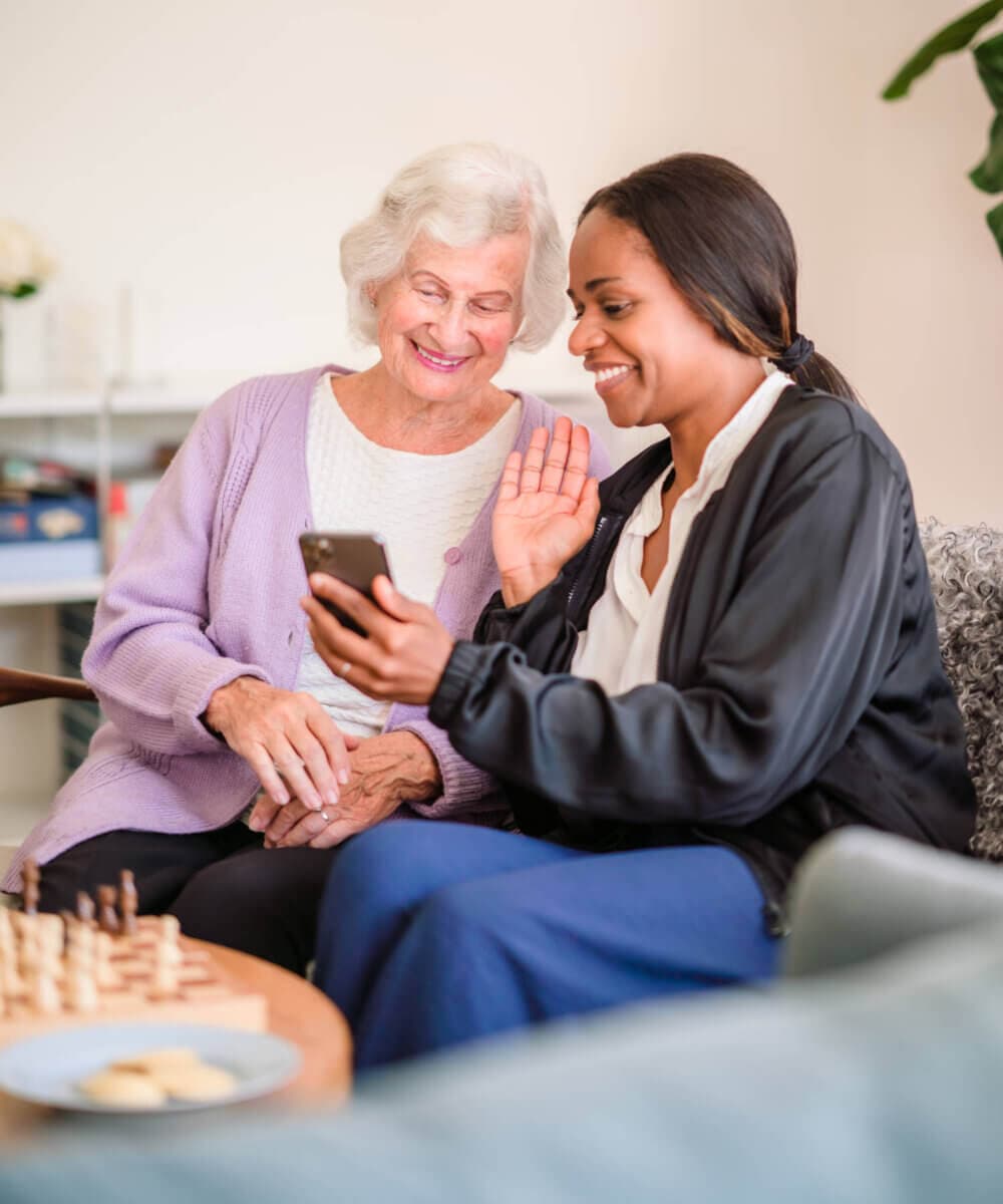 Two women smiling and waving at a phone screen, one elderly with white hair and one younger. A chessboard is in front of them. - Home Instead
