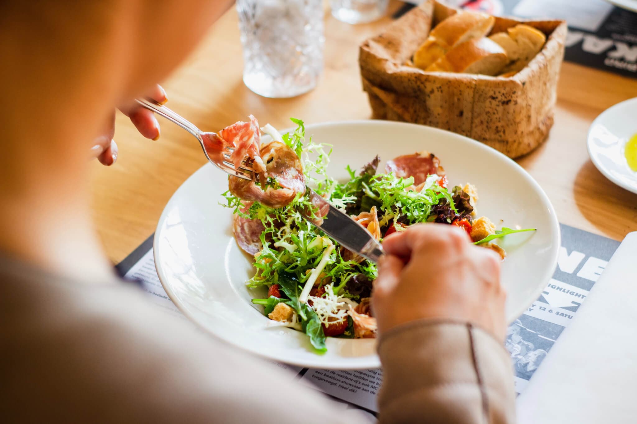 A person is eating a fresh salad with various greens, meats, and toppings at a dining table alongside a basket of bread. - Home Instead