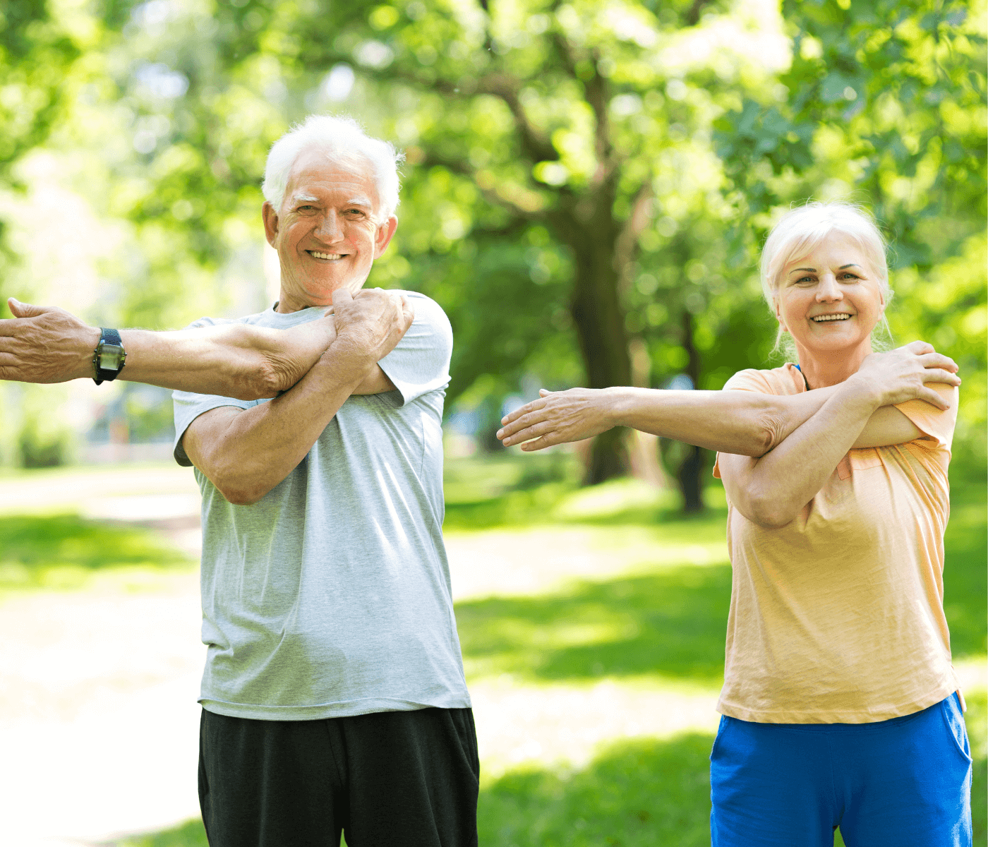 Two smiling elderly individuals exercise in a sunny park, stretching with arms crossed over their chests. - Home Instead