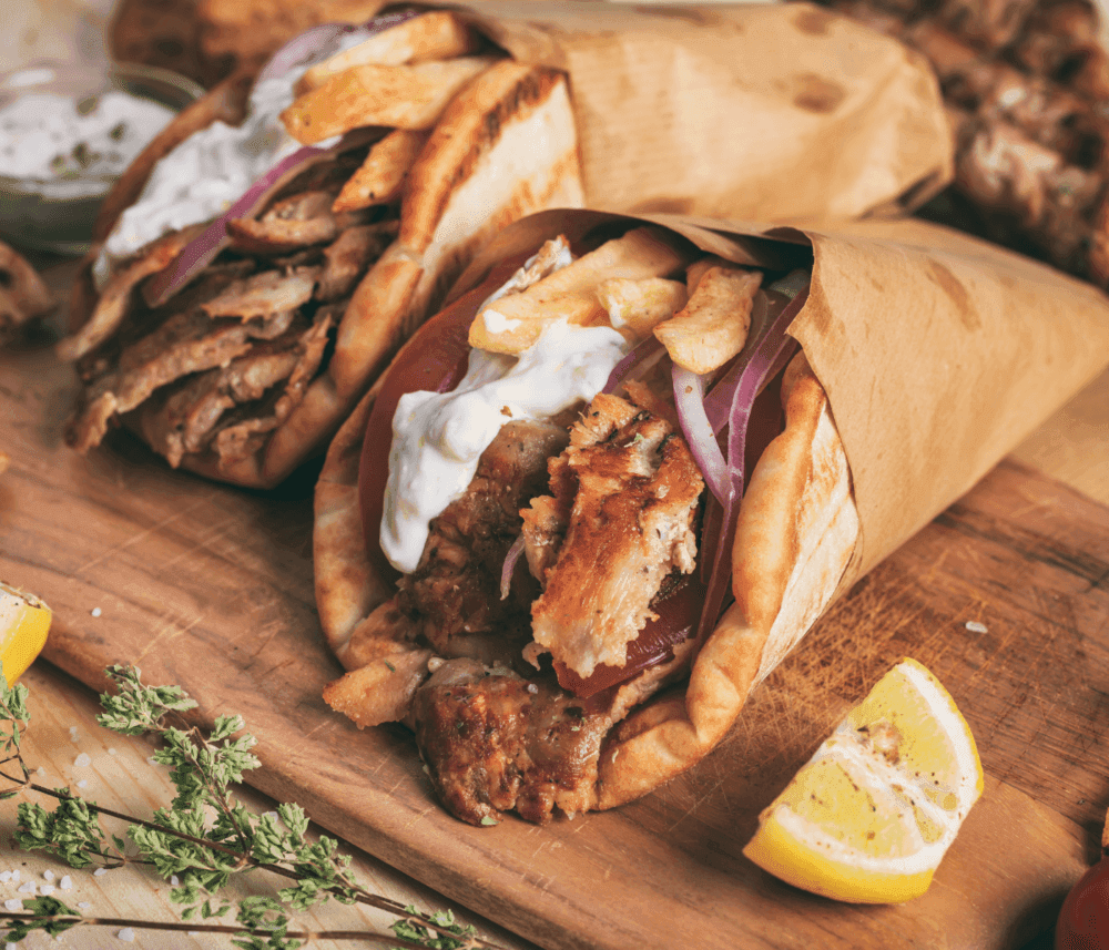 Close-up of two gyros wrapped in paper on a wooden board, garnished with tzatziki sauce, onions, and lemon wedges. - Home Instead