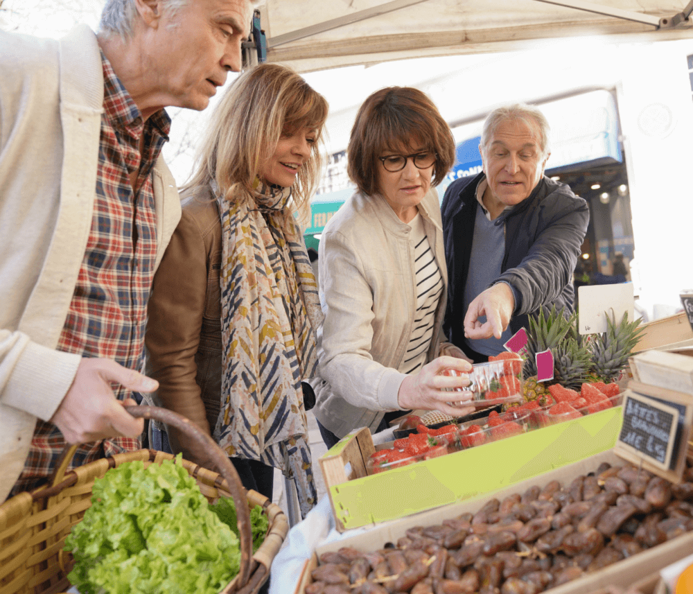 Four people shopping at an outdoor market, selecting strawberries and vegetables, with one holding a basket of lettuce. - Home Instead