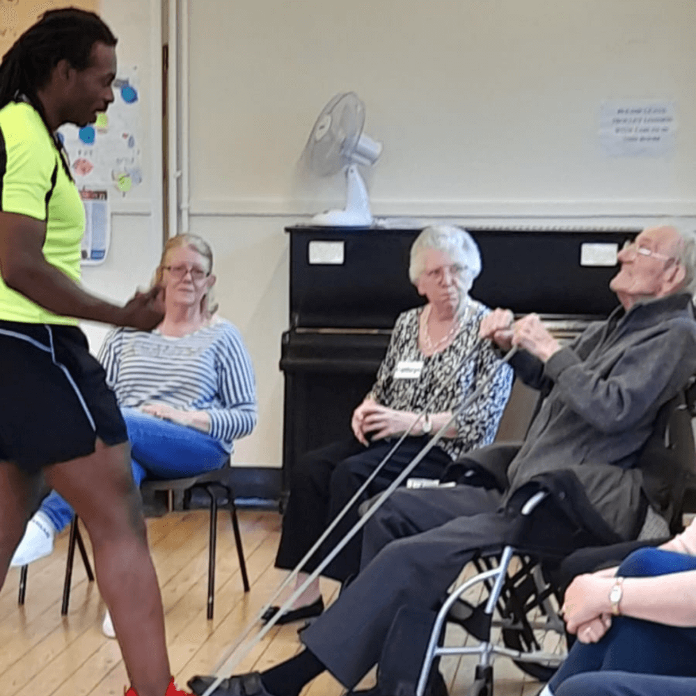 A person in exercise clothes leads a resistance band workout for seated elderly individuals in a community hall. - Home Instead
