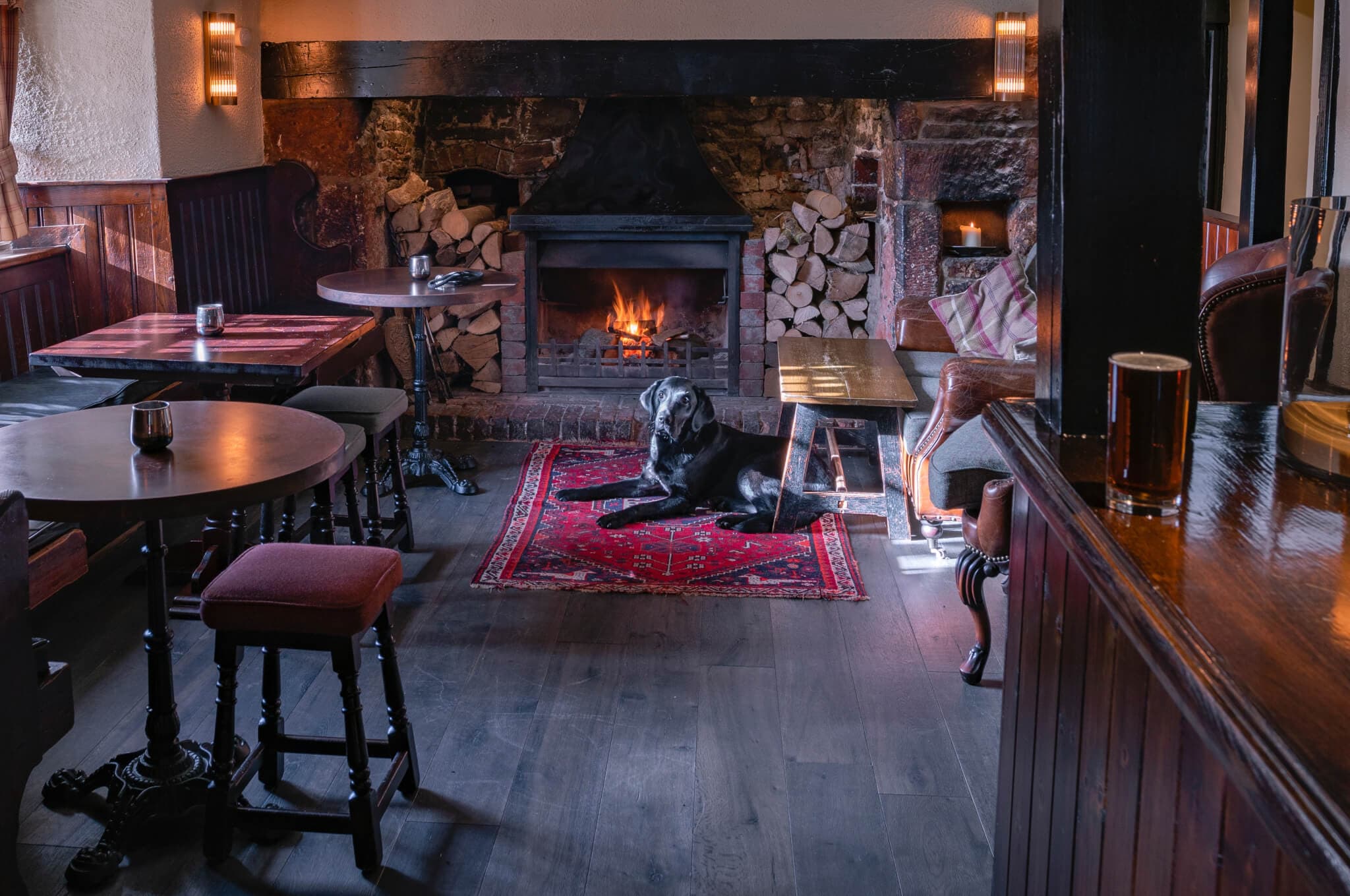 Cozy pub with a lit fireplace, wooden tables, and a black dog resting on a red rug. Log stacks are near the hearth. - Home Instead