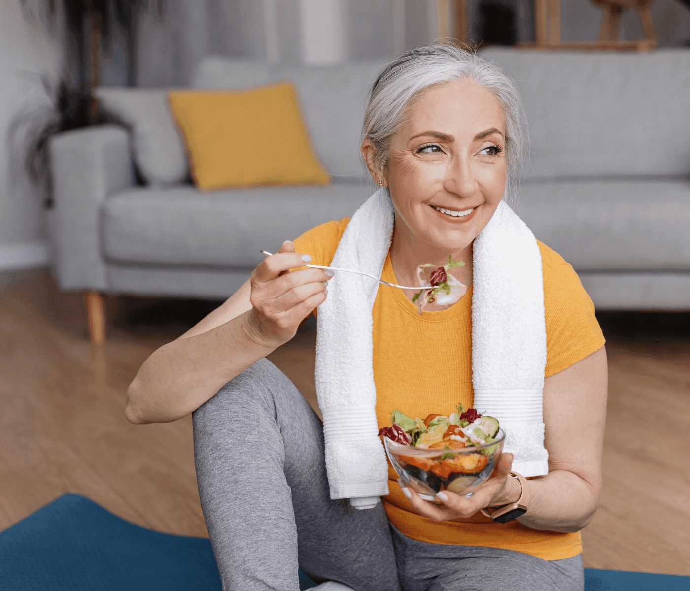 A smiling woman with a towel around her neck eating a salad, sitting on a yoga mat in a living room. - Home Instead