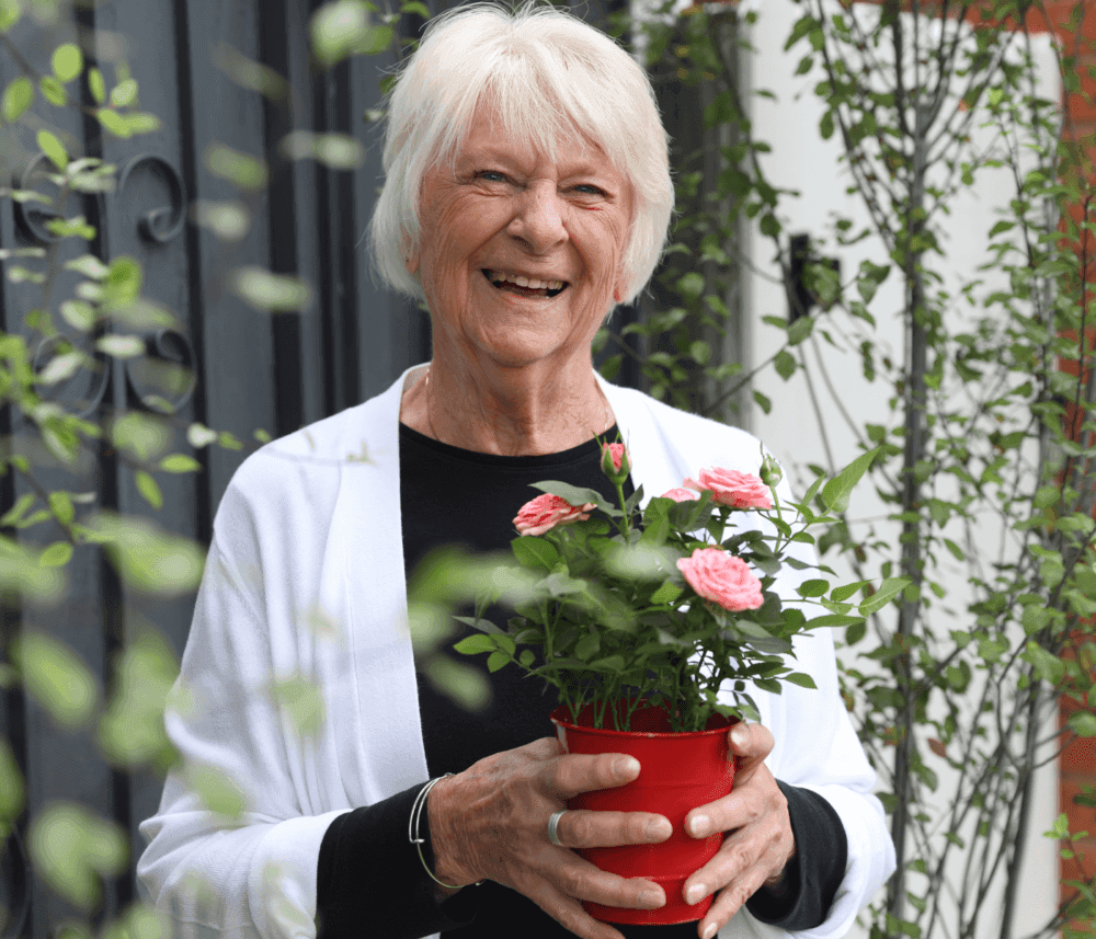 Elderly woman smiles while holding a red pot with pink flowers, standing outdoors with green plants in the background. - Home Instead