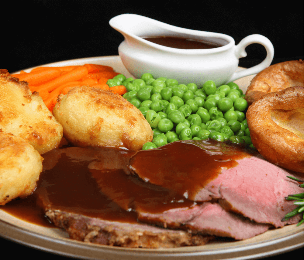 A plate of roast beef with gravy, Yorkshire puddings, roast potatoes, carrots, peas, and a gravy boat in the background. - Home Instead