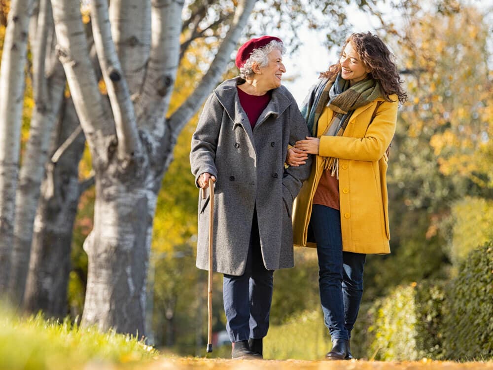 An elderly woman with a cane and a young woman walk arm-in-arm on a tree-lined path in autumn. - Home Instead