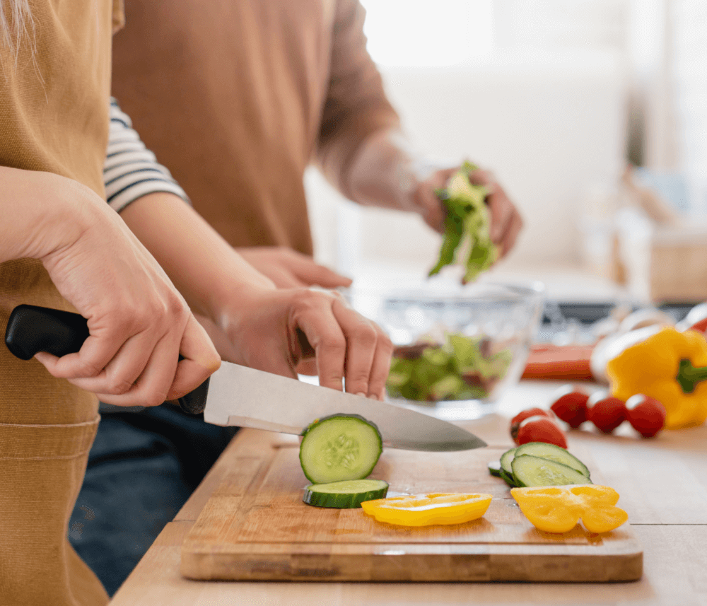 Two people slicing cucumbers and bell peppers on a wooden cutting board, with fresh veggies in the background. - Home Instead