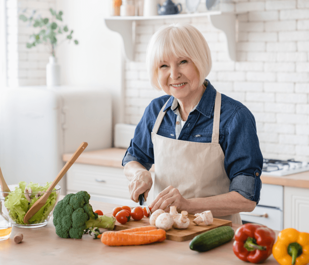 An elderly woman with short white hair chops mushrooms in a bright kitchen with various fresh vegetables on the counter. - Home Instead