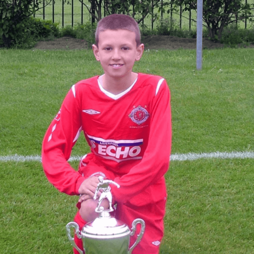 A young boy in a red soccer uniform smiles while holding a trophy on a grassy field. - Home Instead