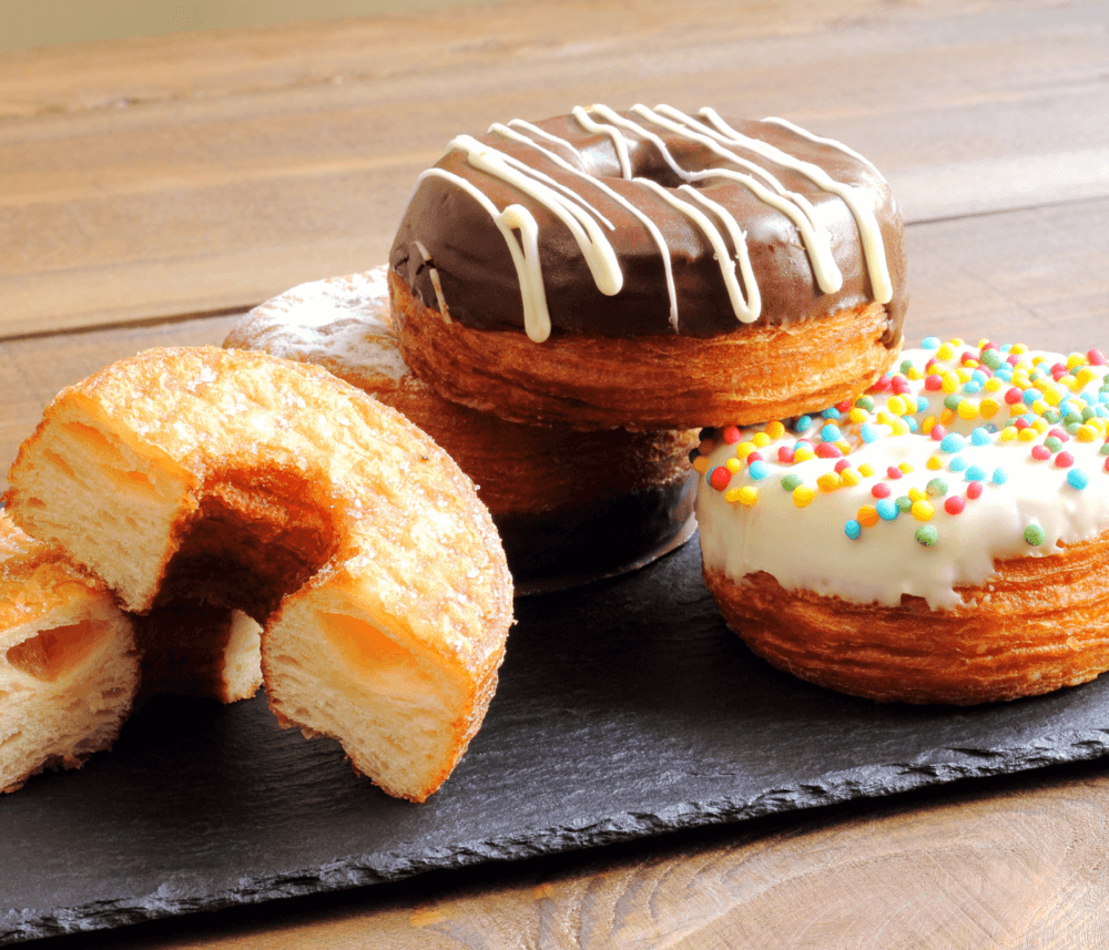 Four cronut pastries on slate: one plain, one with chocolate drizzle, one with rainbow sprinkles, and one split in half. - Home Instead