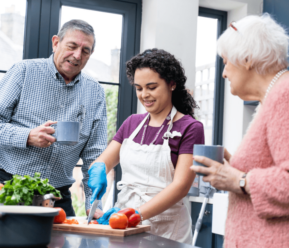 A young woman cuts vegetables while two elderly people watch and drink from mugs in a bright kitchen. - Home Instead