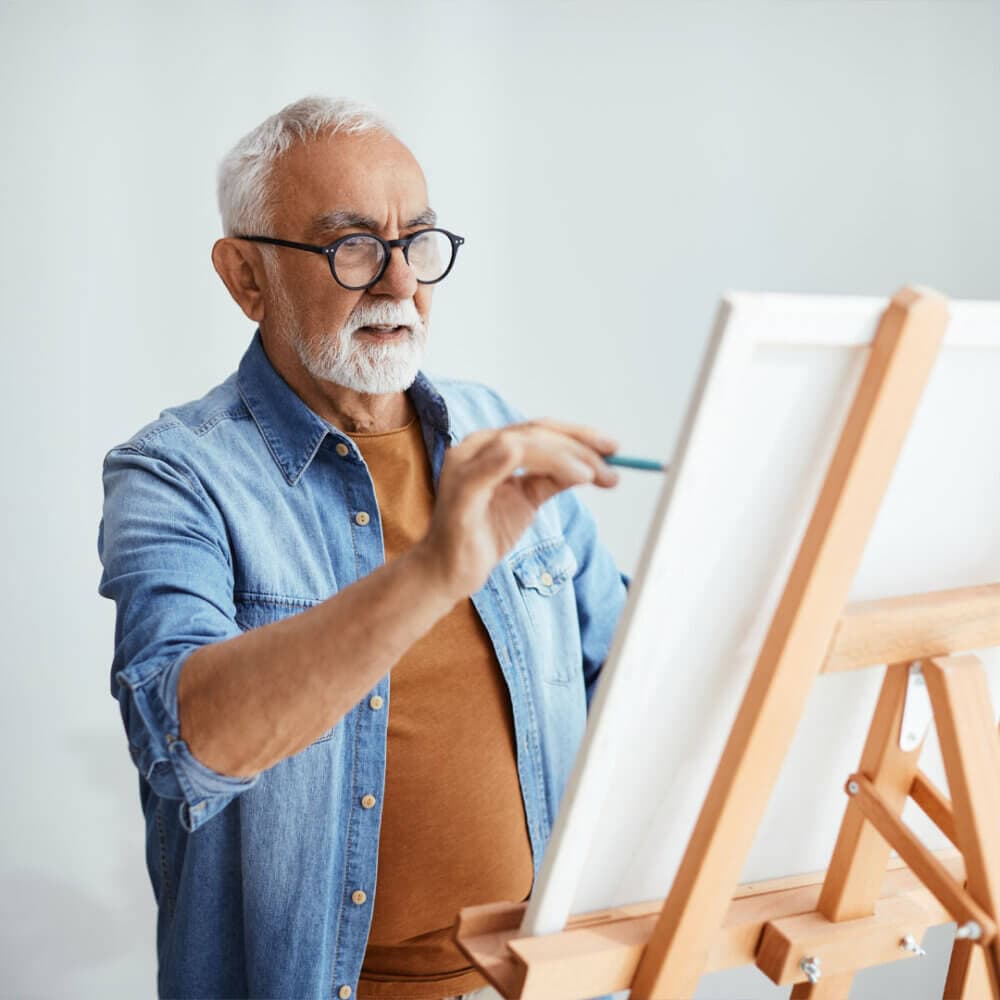 An elderly man with glasses and a denim shirt is painting on a canvas on an easel. - Home Instead