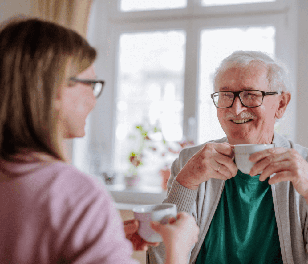 An elderly man and a young woman smiling and drinking tea together at a sunlit table by the window. - Home Instead