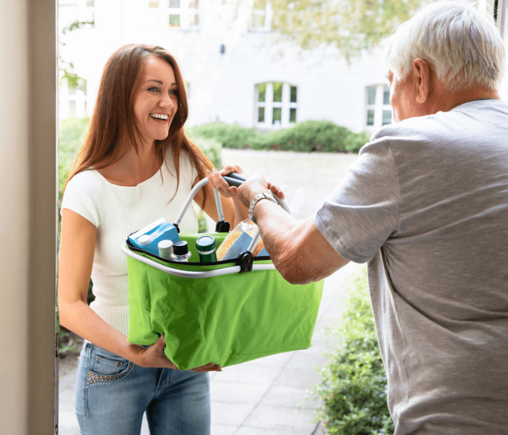 A smiling woman hands a green basket of groceries to an elderly man at a doorstep. - Home Instead