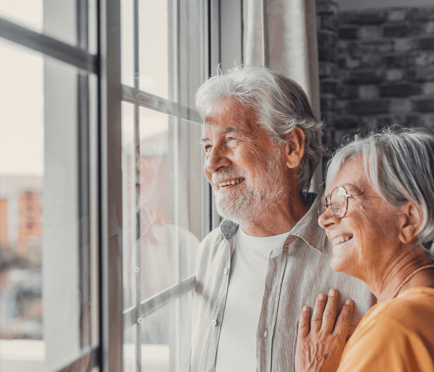 Elderly couple smiling and looking out a window, standing close together in a warmly lit room. - Home Instead