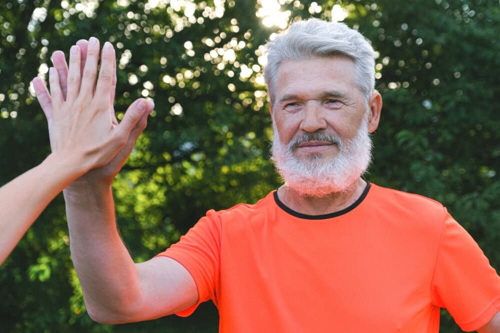 Older man with gray beard and orange shirt high-fives someone outdoors, greenery in the background. - Home Instead