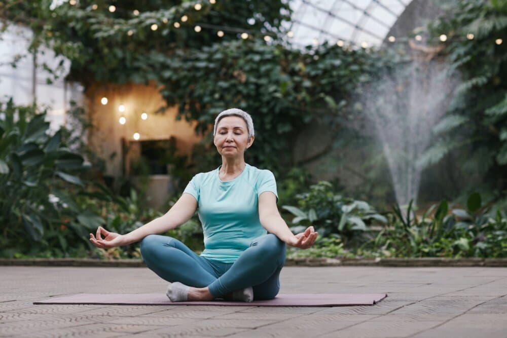A woman meditates in a lotus pose on a yoga mat in a lush, indoor garden with string lights and a fountain in the background. - Home Instead