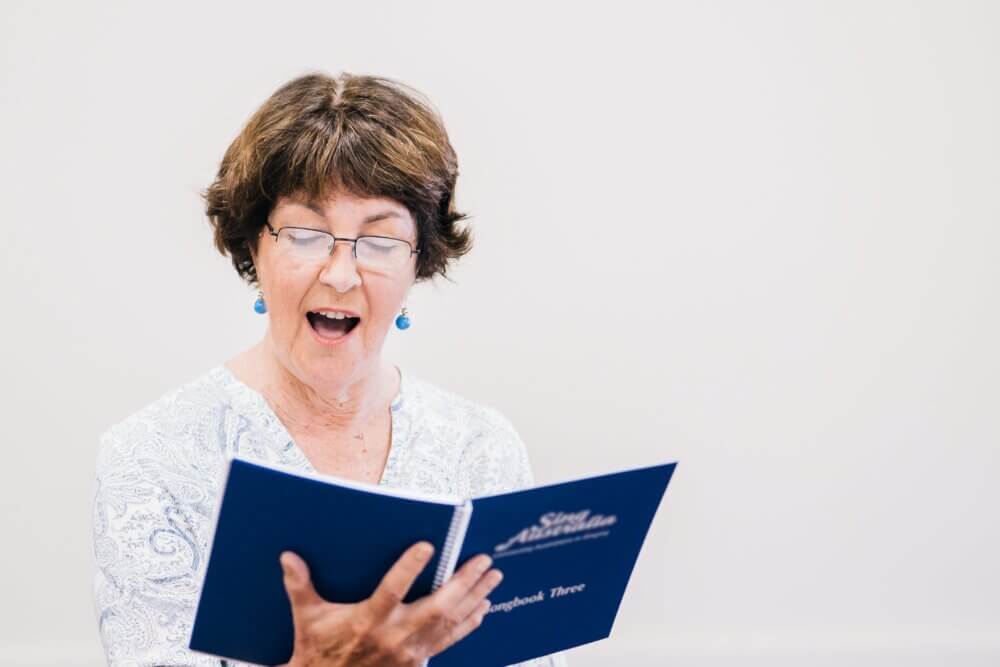 A woman with short brown hair reads and sings from a blue book while wearing glasses and a white patterned blouse. - Home Instead