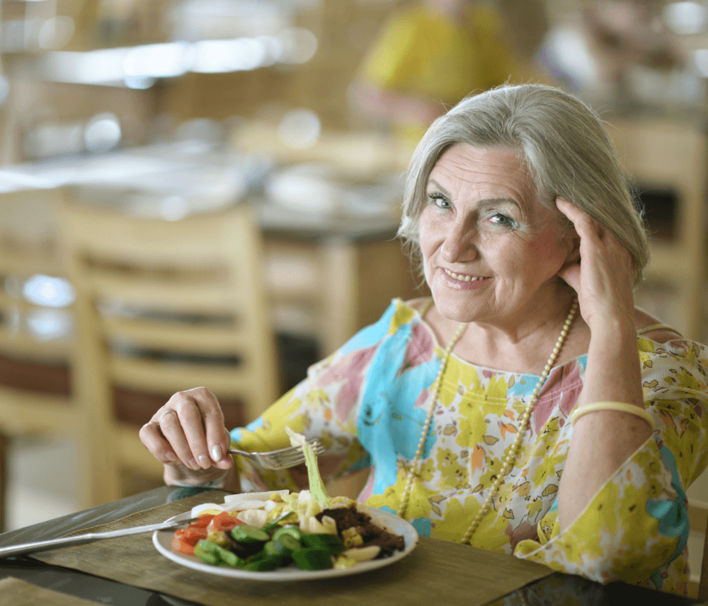 Elderly woman with gray hair and a colorful blouse smiles while eating a meal at a restaurant. - Home Instead
