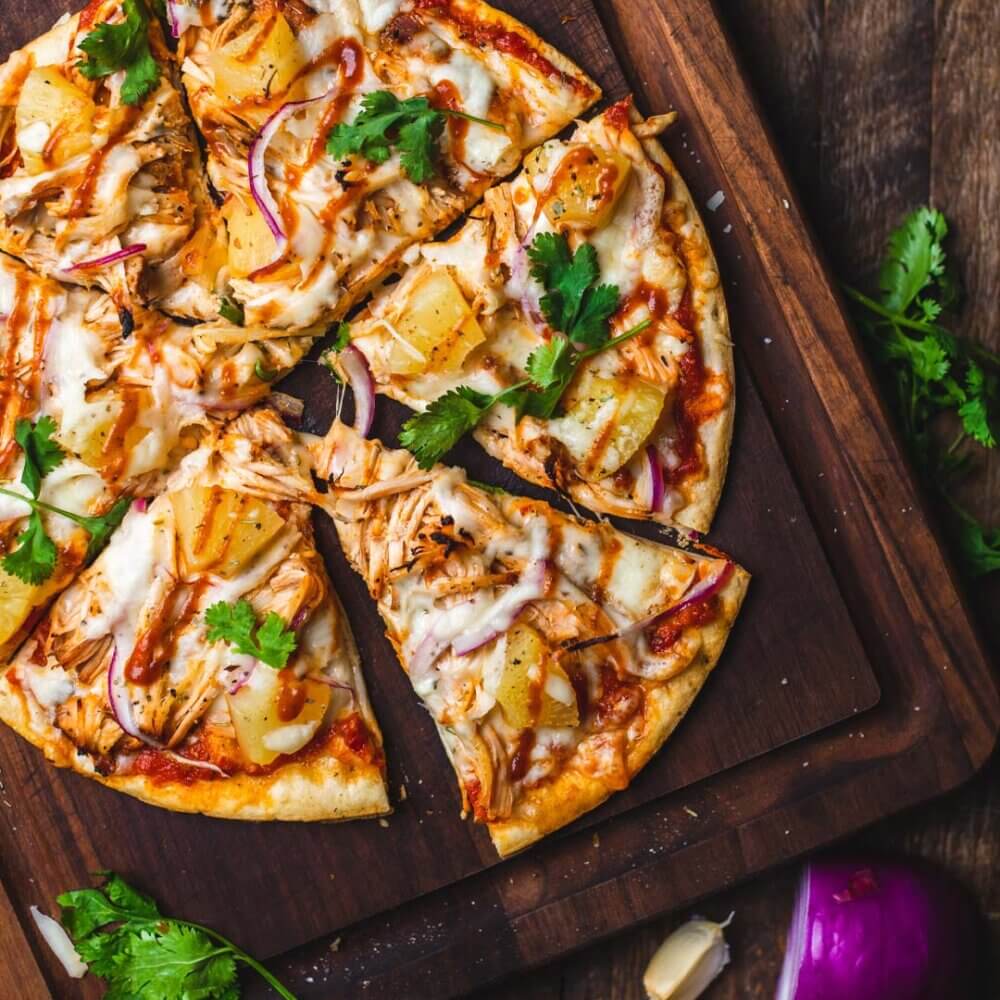 A delicious pizza topped with cheese, pineapple, red onion, cilantro, and barbecue sauce, served on a wooden board. - Home Instead