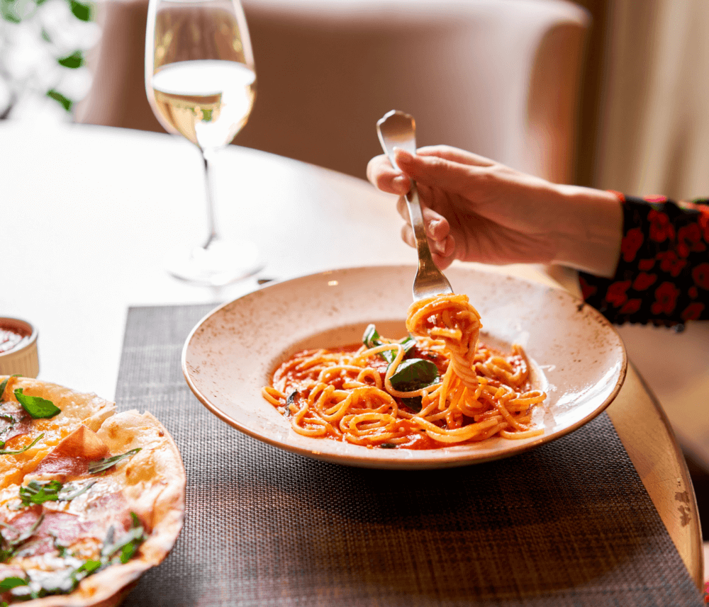 A hand twirls spaghetti with tomato sauce on a fork. A glass of white wine and a pizza are on the table. - Home Instead