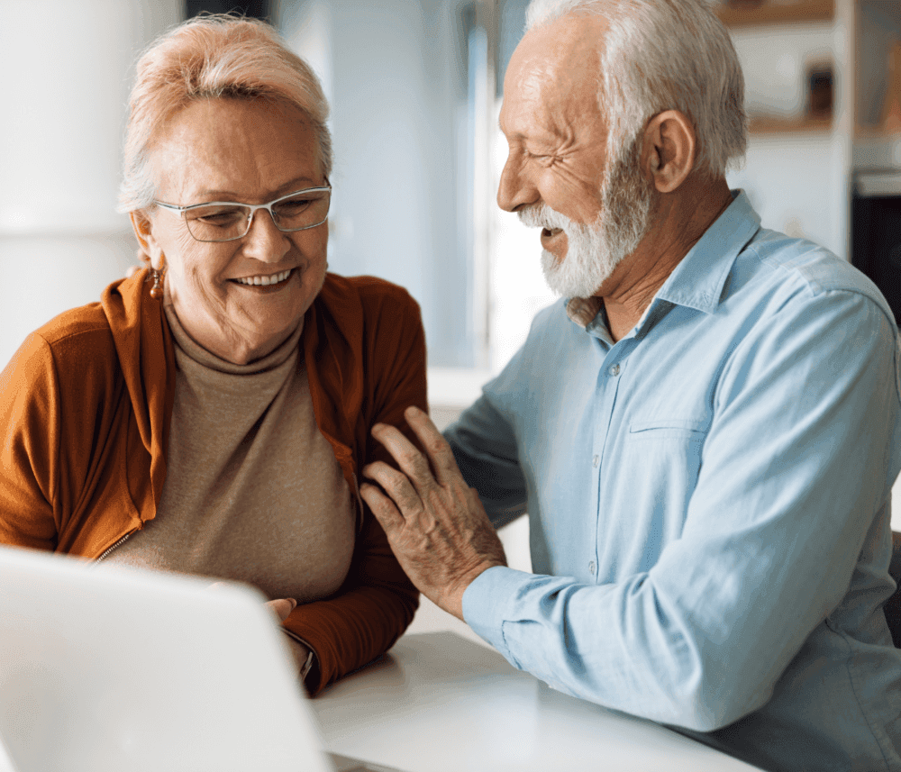 An elderly couple smiling and laughing together while looking at a computer screen in a bright room. - Home Instead